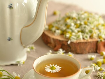 Tasse Kamillentee mit schwimmender Kamillenblüte neben einer Porzellan-Teekanne und frischen Kamillenblüten auf einer Holzscheibe.